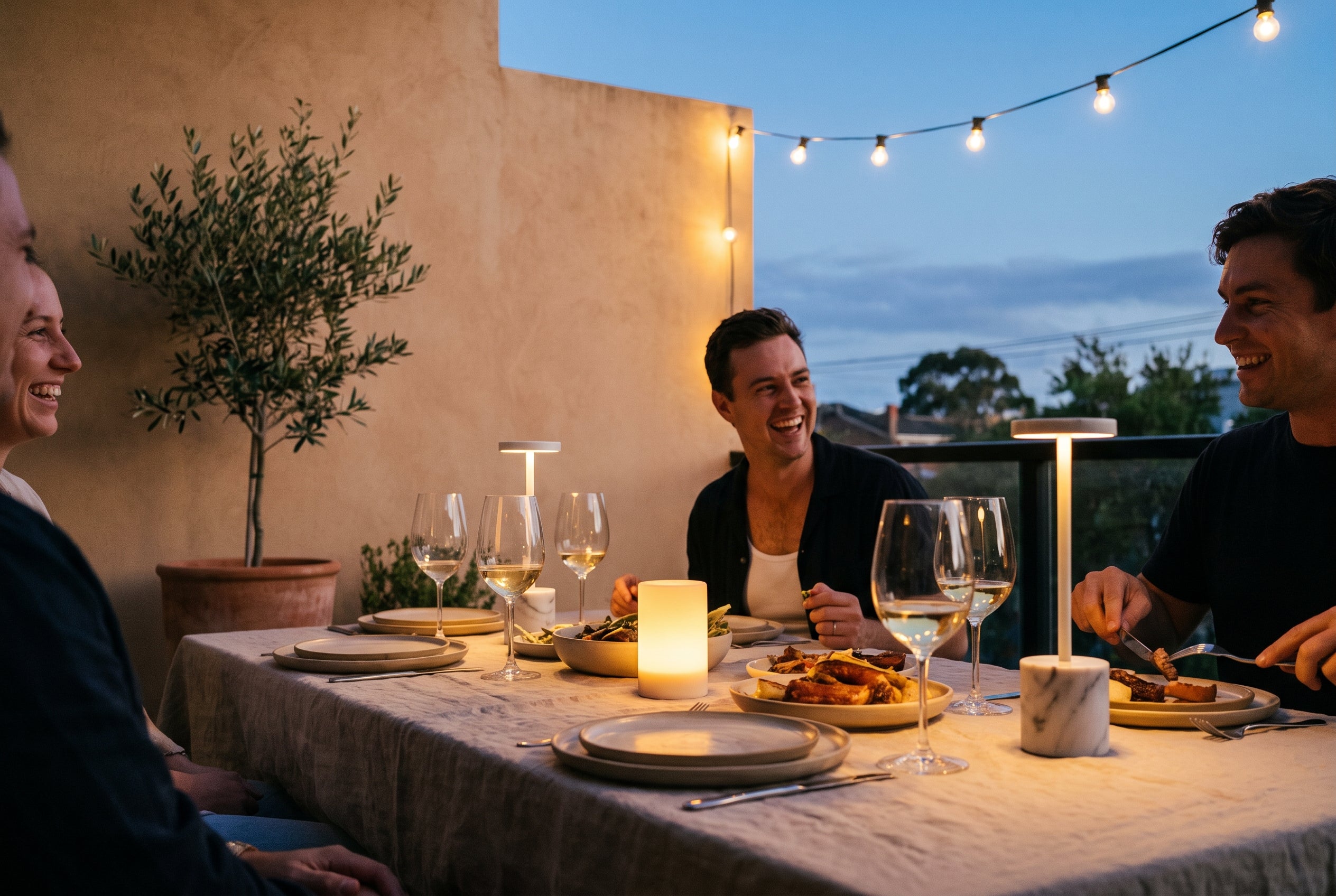 Friends at an outdoor dinner lit by cordless Las Noche lamps at dusk