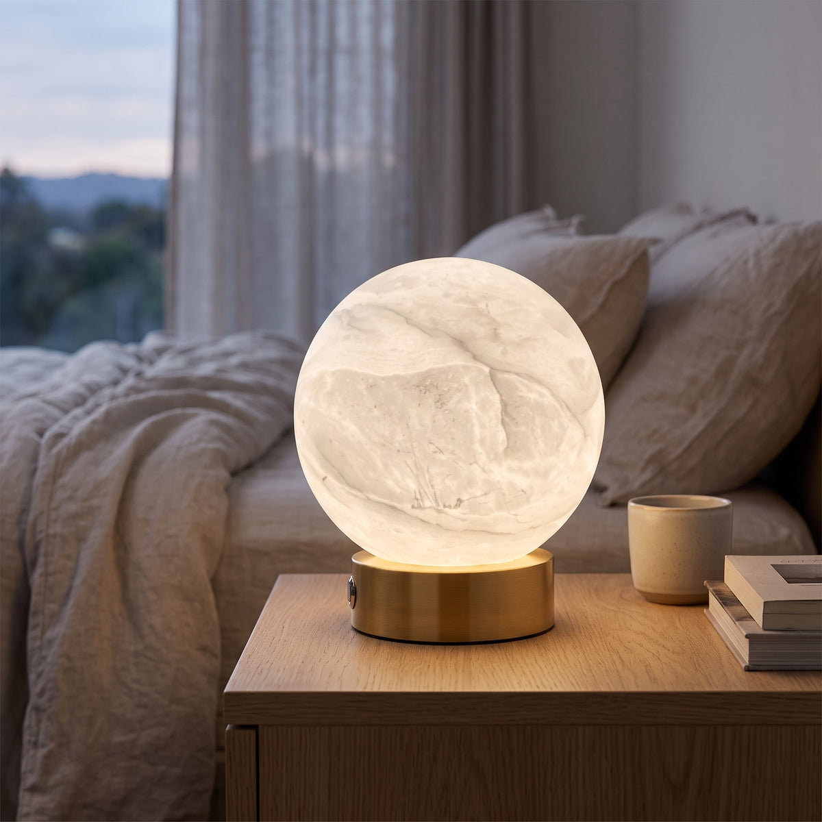 Luna white alabaster sphere lamp on a brass base glowing on a bedside table at dusk