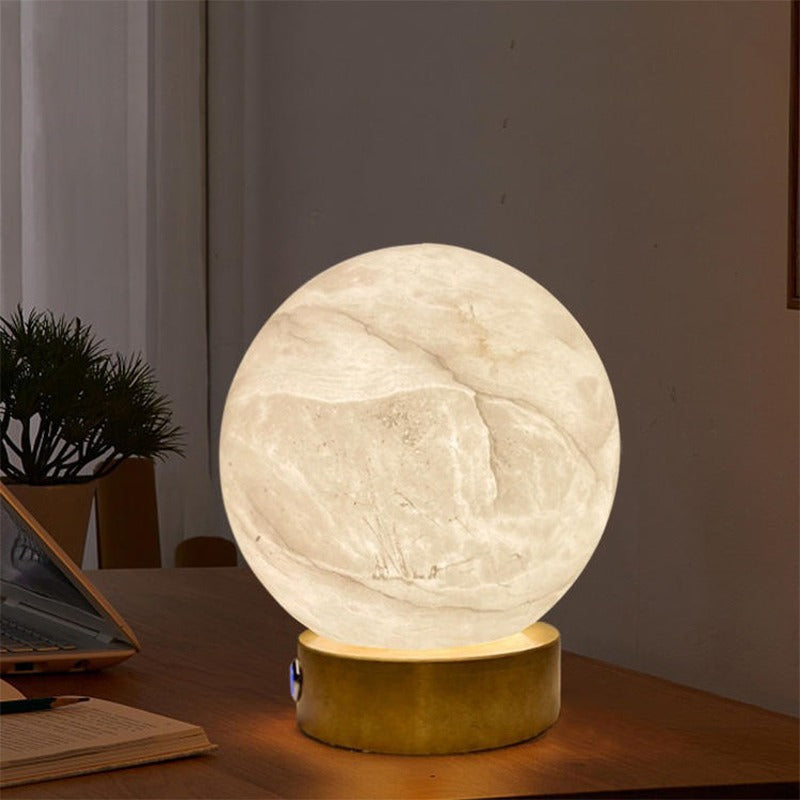 Luna rechargeable alabaster orb lamp lit on a wooden desk next to a laptop and a plant in the evening