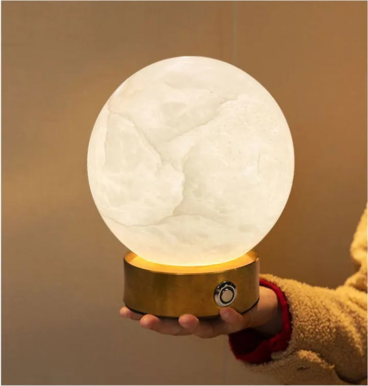 Hand holding the Luna portable alabaster orb lamp, showing its compact size and the button on the brass base